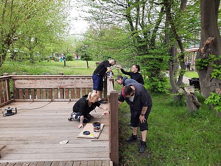Bild zeigt einen Mitarbeiter des Pflegeheimes beim abschleifen des Holzsteges und 4 andere Mitarbeitende beim neu aufbringen der Schutzlasur
