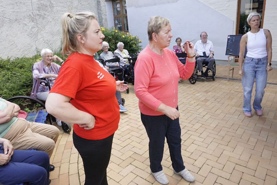 Bild zeigt einen weiblichen Tagesgast beim Darts spielen. Links steht eine Mitarbeiterin der Tagespflege in roter Dienstkleidung und beobachtet die Frau beim Pfeil werfen.