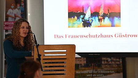 Bild zeigt Frau Ronja Jäger, Einrichtungsleiterin des Frauenschutzhauses in Güstrow, am Rednerpult stehend und zu den Stadtvertretern in Bützow sprechen. Im Hintergrund ist eine Leinwand zu sehen, auf die ein Bild (gemalt Segelboote die einen Hafen verlassen bzw. ansteuern - nur schemenhaft zu erkennen) projiziert wird. Darunter der Schriftzug "Das Frauenschutzhaus Güstrow"