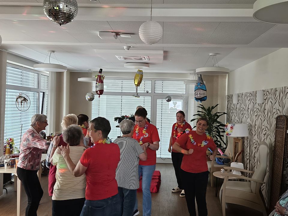 Bild zeigt eine ausgelassene Stimmung im hellen Gemeinschaftsraum während einer 'Roland-Kaiser-Party' in der DRK Tagespflege. Mehrere Senioren und jüngere Pflegekräfte, einige davon in roten DRK T-Shirts und mit bunten Blumenketten geschmückt, tanzen und singen. Im Hintergrund sind Fenster mit Jalousien, ein glänzender Party-Luftballon in Flaschenform und eine Discokugel an der Decke zu erkennen. Die tanzenden Personen bilden eine lockere Gruppe in der Mitte des Raumes.