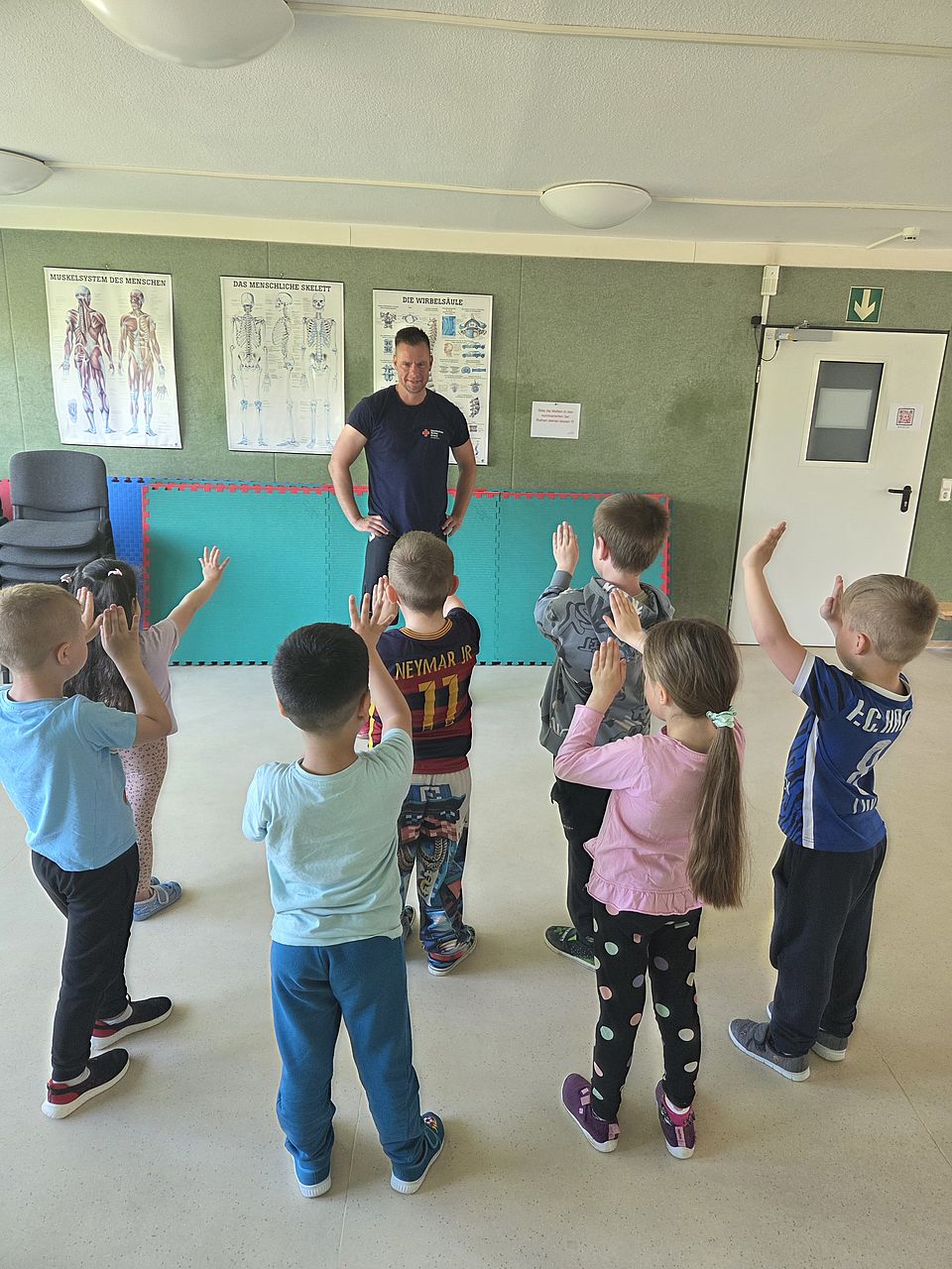 Bild zeigt eine Gruppe von kleinen Kindern mit dem Rücken zur Kamera stehend und machen die Bewegungen des Übungsleiters nach