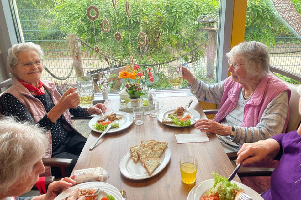 Bild zeigt: Vier ältere Damen sitzen fröhlich an einem Tisch am Fenster und feiern ein Fest. Zwei der Frauen (links und rechts im Bild) stoßen mit vollen Bierkrügen an und lächeln. Auf dem Tisch stehen Teller mit Mahlzeiten (Fleisch, Salat), Beilagen (Brot/Fladenbrot) und frische Blumen. Die Damen tragen festliche Kleidung, unter anderem eine rosa Weste. Im Hintergrund sind durch das Fenster Gartendekoration und Bäume sichtbar. Die Szene zeigt ein gemütliches Beisammensein und Feier in der Tagespflege.