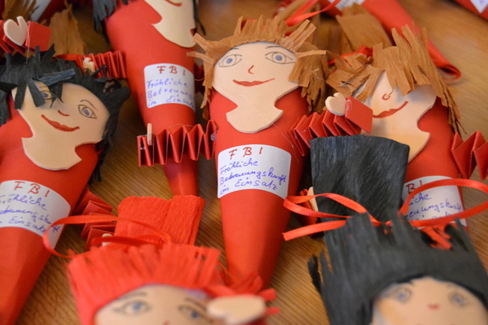 Weiterbildung Bild 4 Bild zeigt die selbstgebastelten, kleinen Schultüten in rotem Papier mit aus Moosgummi geschnittenen Gesichtern, bemalt mit Augen, Nase und Mund sowie aus Krepppapier aufgeklebten Haaren. Auf jeder Tüte klebt ein weißes Schild mit der Aufschrift in roten Buchstaben F B I darunter in blauer Schrift die Erklärung: Fröhliche Betreuungskraft im Einsatz.