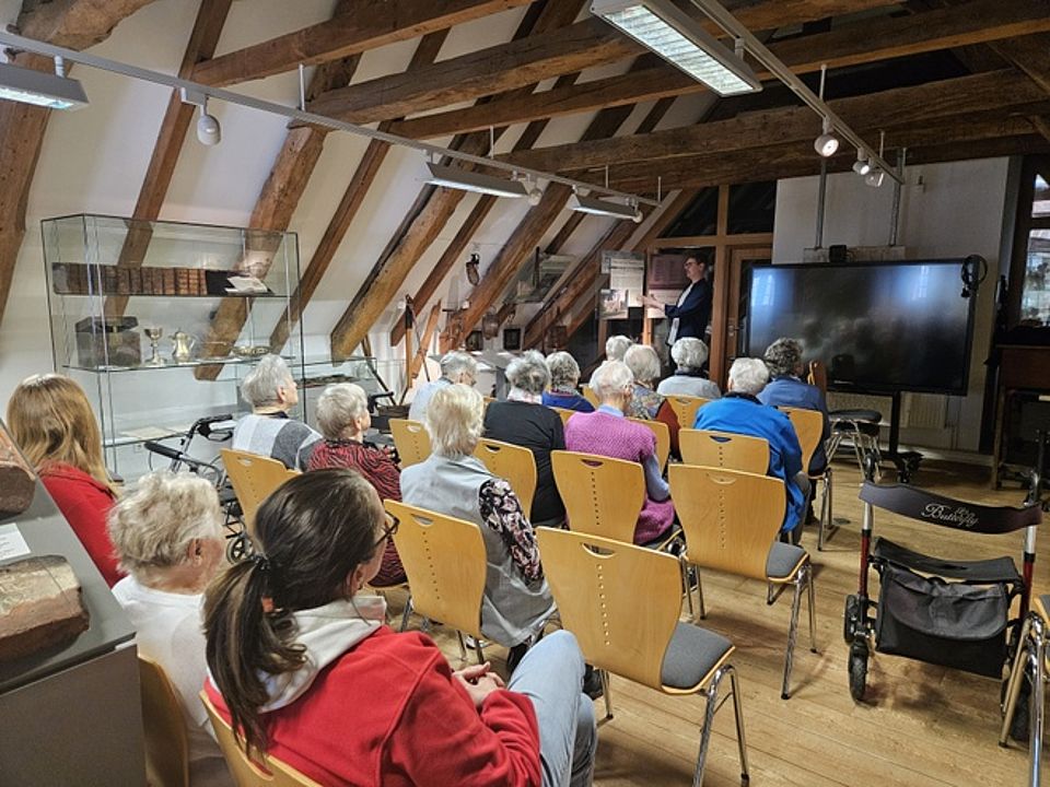 Bild zeigt einen Blick in den Vorführraum des Museums mit großem Flachbildschirm und Stuhlreihen davor. Auf diesen Stühlen sitzen Besucher der Tagespflege und hören den Erklärungen eines Museumsmitarbeiters gespannt zu.