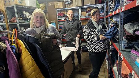 Bild zeigt eine Blick in die Güstrower DRK Kleiderkammer. Es sind gefüllte Regale und Kleiderständer zu sehen. Die Mitarbeitenden der Kleiderkammer stehen mit im Raum und halten Kleidungsstücke in den Händen.