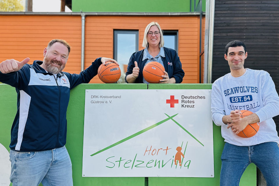 Bild zeigt:  Anja Wulff, Einrichtungsleitung DRK-Hort Stelzenvilla (M.), Tobias Hahn vom Verein „Basketball macht Schule“ (l.) und Trainer Abdulaziz Bayrak vor dem Gebäude des Hortes Stelzenvilla. Jeder hält einen Basketball in der Hand Alle haben lachende und freundliche Gesichter