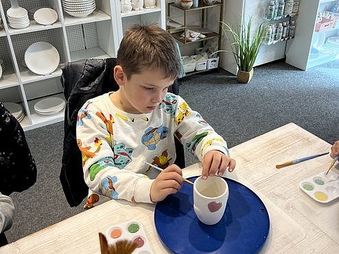 Bild zeigt eine Jungen an einem Tisch sitzend. Vor ihm steht auf einer blauen drehbaren Unterlage ein Keramikbecher. Der Junge trägt einen weißen Pullover mit bunten Motiven. Neben der Unterlage steht ein Malkasten mit mit verschiedenen Farben. Im Hintergrund ist eine Große Regalwand mit ganz vielen verschiedenen Keramikschüsseln -teller und -tassen. -