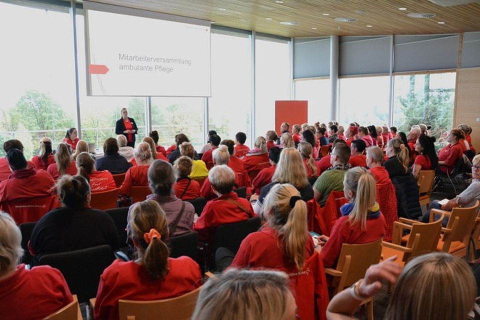 Rund 100 Mitarbeitende Bild zeigt einen großteil der Teilnehmenden der Mitarbeiterversammlung der ambulanten Pflege in Stuhlreihen sitzend und den Begrüßungsworten von Frau Cornelia Heidenreich zuhörend.