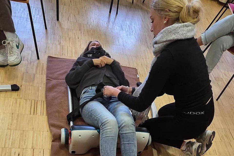 Bild zeigt zwei junge Frauen beim testen eines weiteren Hilfsmittels in der Pflege. Eine der Frauen liegt auf dem Boden und wird an ein Gerät geschnallt. Die zweite Frau schließt die Gurte.