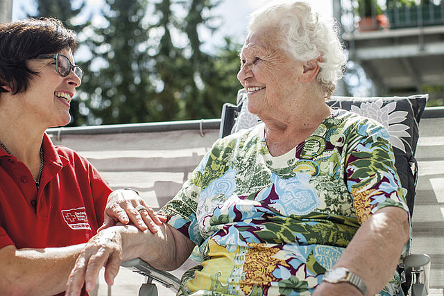 Bild zeigt ein Symbolfoto zum Thema Tagespflege - eine Pflegefachkraft beschäftigt sich mit einer Seniorin Hände haltend und lachend