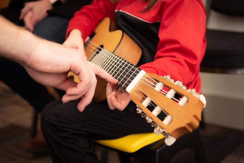 Bild zeigt den Gitarrenhals einer von einem Kind gehaltenen Gitarre. Eine erwachsene Hand kommt von links ins Bild und drückt mit dem Zeigefinger die hohe e-Saite im dritten Bund, so dass das Kind weiß, wie es seinen Finger setzen muss.