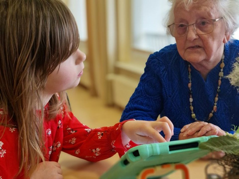 Bild 1 Bild zeigt ein junges Mädchen aus der dritten Klasse mit einer Seniorin am Tisch sitzend und ein digitales Tablet erklöärend. Das kleine Mädchen trägt eine rote Bluse mit kleinen Blumen drauf, die Seniorin trägt einen blauen Pullover mit einer (vermutlich) Bernsteinkette.