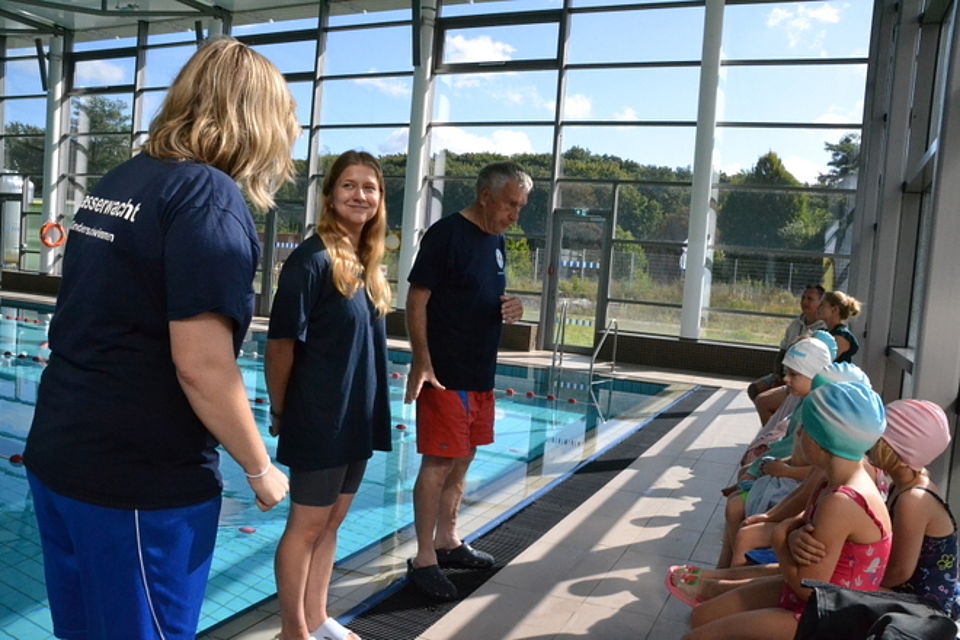 Bild zeigt die Schwimmlehrer stehen vor einer Gruppe Kita-Kinder, welche sitzend den Einweisungen der Schwimmlehrer zuhören. Im Hintergrund ist das Trainingsbecken zu sehen und ein Blick durch die große Fensterfront.