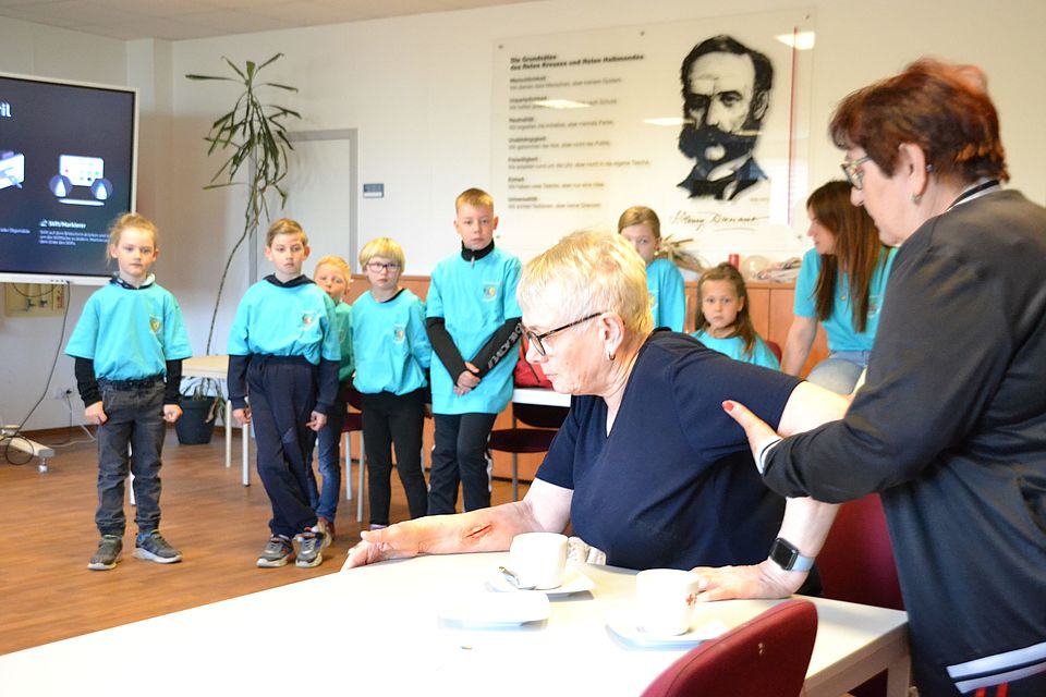 Bild zeigt im Vordergrund eine verletzte Person am Tisch sitzend. Im Hintergrund steht eine Mannschaft junger JRKler und berät wie und in welcher Reihenfolge jetzt zu helfen ist.