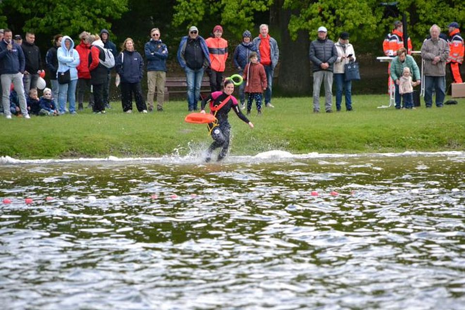 Wasserrettung Bild zeigt Frau Hein, welche mit Rettungsleine und orangefarbener Rettungsboje in den Inselsee läuft. Sie trägt einen Neoprenanzug mit roten Schulterpolstern. Im Hintergrund stehen über 20 Personen, die sich die Demonstration interessiert anschauen.