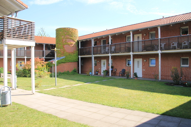 Bild zeigt die Ansicht des Innenhofes der Anlage in Krakow. Die einzelnen Zugänge zu den Wohneinheiten sowie der Laubengang sind zu sehen. in der Mitte ist eine große, schön gepflegte Rasenfläche. Im Hintergrund ist ein haushoher Turm zu sehen, der mit wildem Wein bewachsen ist.