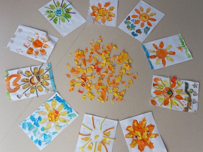 Bild zeigt eine auf dem Fußboden liegende Collage. Es liegen 10 A4 Blätter, die sich mit den oberen Ecken berühren und somit einen Kreis bilden. In der Mitte des Kreises liegen Tonpapier-Schnipsel in den Farben Gelb, Rot und Orange. die Bilder zeigen zeigen Sonnen- manche gemalt mit Tusche, andere geklebt mit Schnipseln aus Tonpapier.