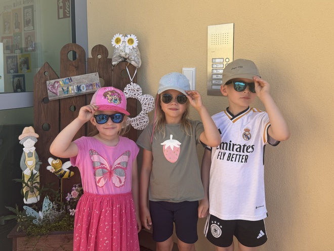 Bild zeigt 3 Kinder vor dem Haupteingang der Kita mit sommerlicher Kleidung - T-Shirt und Shorts, das Mädchen trägt ein Pinkfarbenes Sommerkleid mit Schmetterlingmotiv,  alle Kinder tragen ein Basecap und eine Sonnenbrille