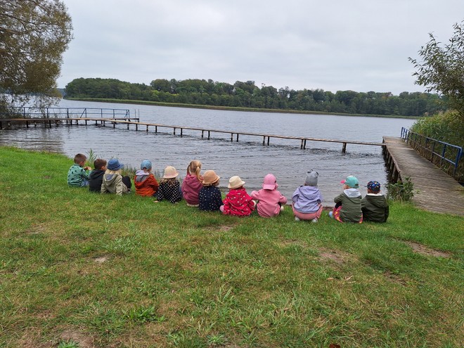 Bild zeigt: Zwölf Kleinkinder, alle mit Mützen und Jacken bekleidet, sitzen auf einer Naturexkursion nebeneinander auf einer grasbewachsenen Uferböschung und blicken auf einen See. Die Kinder sind von hinten zu sehen. Auf dem ruhigen See ist eine lange, hölzerne Steganlage zu erkennen, die ins Wasser ragt. Das Ufer ist von Schilf und Bäumen gesäumt. Die Szene vermittelt den Eindruck eines ruhigen Ausflugs an einem See an einem kühleren Tag.