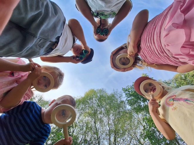 Bild zeigt sechs Kinder, welche durch eine Lupe auf den Boden schauen. Das Bild ist vom Boden in Richtung Himmel aufgenommen, so dass man die Gesichter der Kinder durch die Lupe sehen kann.