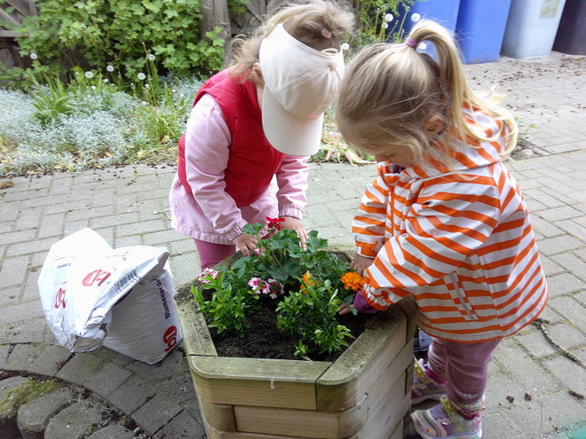 Bild zeigt: Zwei kleine Mädchen pflanzen Blumen in einem hölzernen Blumenkübel auf einem gepflasterten Weg. Das Mädchen links trägt eine rosafarbene Weste über einem hellen Oberteil und eine beige Schirmmütze. Es beugt sich über den Kübel und arrangiert Pflanzen mit weißen und roten Blüten. Das Mädchen rechts trägt eine auffällige orange-weiß gestreifte Jacke und hat blondes Haar zu einem Pferdeschwanz gebunden. Es ist ebenfalls dabei, eine orangefarbene Blume in die Erde zu setzen. Neben dem Kübel liegt ein aufgerissener Sack Blumenerde. Im Hintergrund sind ein Gartenbeet und ein blauer Mülleimer zu sehen, was die Szene als Outdoor-Aktivität im Kindergarten kennzeichnet.
