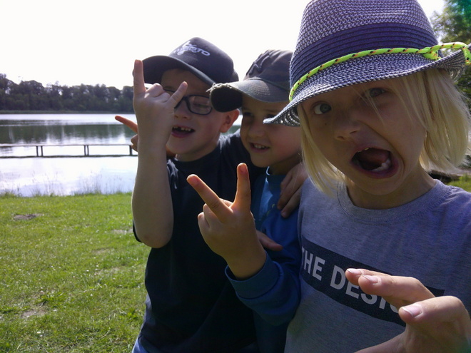 Foto-Rallye Bild zeigt: Nahaufnahme von drei fröhlichen Kindern, die Grimassen schneiden und Handzeichen geben, während sie vor einem See stehen. Das Kind links trägt eine schwarze Kappe und Brille und macht das "Pommesgabel"-Zeichen (Rock-Geste). Das Kind in der Mitte lächelt, trägt eine dunkle Kappe und macht das Friedenszeichen (Victory-Zeichen) mit der Hand. Das Kind rechts, mit blondem Haar und einem grauen Sonnenhut mit neongrünem Band, reißt den Mund auf und macht eine Grimasse, während es auf den Betrachter zeigt. Im Hintergrund sind eine grüne Wiese, ein See und ein kleiner Holzsteg zu sehen.