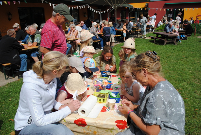 Bild zeigt: Viele Erwachsene und Kindern sitzend an Tischen im Freien. Der Tisch im Vordergrund ist mit einer gelben Tischdecke, Bemalungen und einer Küchenpapierrolle, sowie mit ganz vielen anderen Bastelmaterialien bedeckt. Zwei Frauen und mehrere Kinder mit sommerlichen Hüten und Mützen sind vertieft in die Bastelaktivitäten (Malen und Kneten), umgeben von bunten Behältern mit Bastelmaterial. Im Hintergrund sind zahlreiche weitere Personen an Bierbänken und Picknicktischen auf einer Wiese versammelt, umgeben von Gebäuden mit bunten Fassaden (Orange und Rot). Über den Köpfen der Menge hängen bunte Wimpelketten.