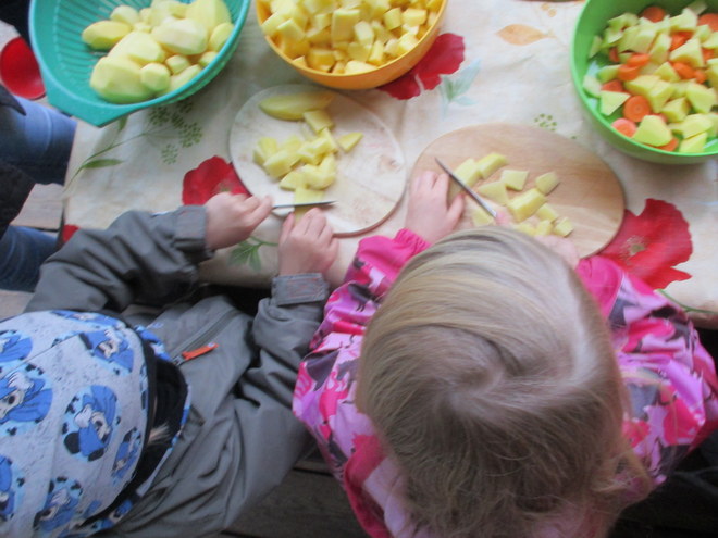 Bild zeigt: Aufsicht auf zwei Kinder, die auf einem Tisch Kartoffeln schneiden und zum kochen vorbereiten. Die Kinder sitzen nebeneinander. Das blonde Kind rechts trägt ein pink-gemustertes Oberteil und hält ein Messer, um Kartoffelstücke auf einem kleinen runden Holzbrett zu halbieren. Das andere Kind trägt eine graue Jacke und eine Mütze mit Muster und schneidet ebenfalls Kartoffeln auf einem hellen runden Holzbrett. Auf dem Tisch stehen drei bunte Schüsseln (türkis, gelb, grün) gefüllt mit geschälten, gewürfelten Kartoffeln und gewürfeltem Gemüse (Karotten). Die Szene spielt auf einer Tischdecke mit rotem Blumenmuster.