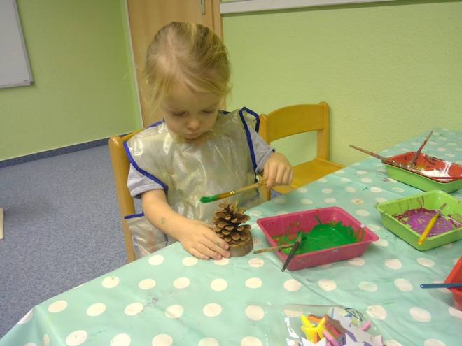Bild zeigt: Ein kleines, blondes Mädchen sitzt an einem Tisch in einem hellgrünen Raum und bemalt einen Tannenzapfen. Das Mädchen trägt einen durchsichtigen Malschurz und konzentriert sich auf den großen Tannenzapfen, den es mit einem Pinsel hält. Vor ihm stehen mehrere flache Plastikschalen (z. B. in Pink und Grün) gefüllt mit bunter Farbe, darunter leuchtendes Grün. Der Tisch ist mit einer hellgrünen Wachstuchdecke mit weißen Punkten bedeckt. Die Szene zeigt eine Bastelaktivität im Kindergarten.