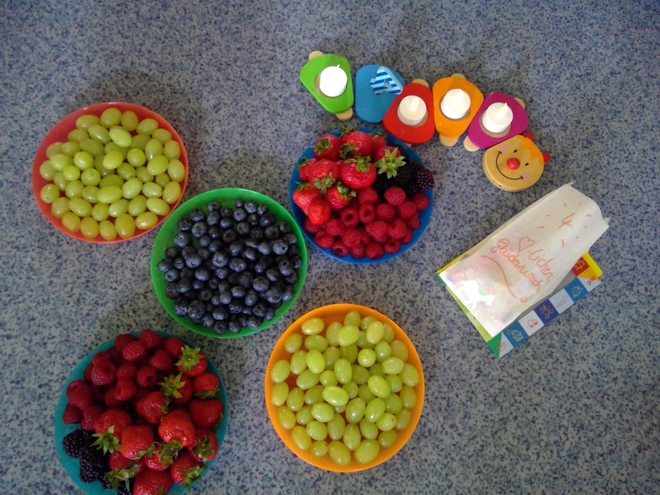 Bild zeigt: Aufsicht auf fünf bunte Schüsseln gefüllt mit frischem Obst für eine Obstpause. Die Schüsseln enthalten grüne Weintrauben (zweimal), Blaubeeren (einmal) sowie eine Mischung aus Erdbeeren, Himbeeren und Brombeeren (zweimal). Die Schalen stehen auf einer blaugrauen, gesprenkelten Oberfläche. Rechts daneben befindet sich eine hölzerne, bunte Dekoration in Form einer Raupe, in deren Segmente Teelichter eingesetzt sind. Ebenfalls ist eine Geschenktüte mit der handgeschriebenen Aufschrift "Herzlichen Glückwunsch" zu sehen.