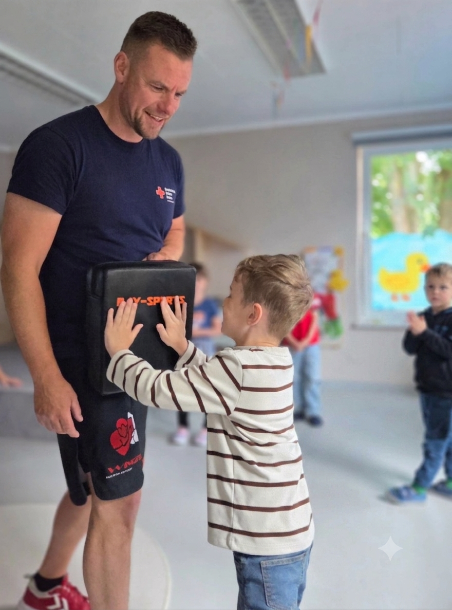 Bild zeigt einen kleinen Jungen, der auf das Schlagpolter, welches der Trainer hält, kraftvoll gegenschlägt. Die gesamte Szene ist im Sportraum der Kita und im Hintergrund sind noch ein paar Kinder unscharf zu sehen.