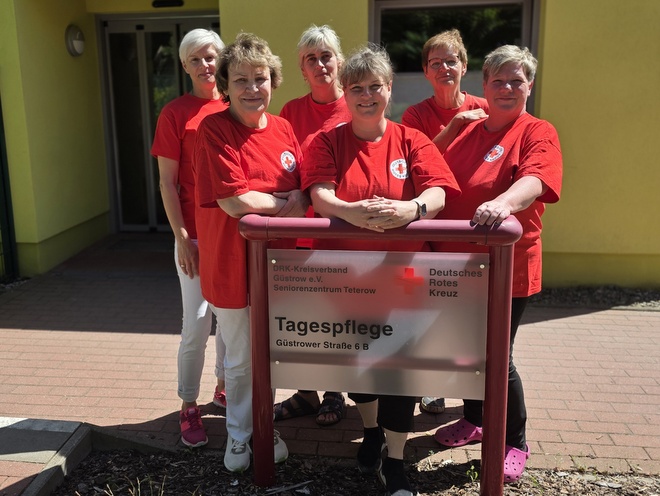 Bild zeigt das Team der DRK Tagespflege in Teterow. Die 6 Mitarbeitenden sind in DRK-roten Shirts fotografiert und stehen vor dem Haupteingang der Tagespflege hinter dem Schildaufsteller, welcher die Anschrift und den Titel der Einrichtung trägt.